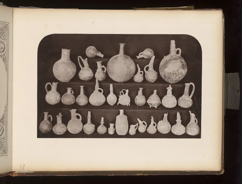 Three rows of small pottery jugs, juglets and flasks set on a dark cloth, photographed in an album.
