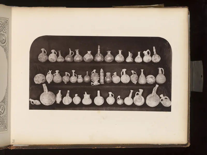 Three rows of small pottery juglets, jugs and round vessels laid out on a dark board, mounted on an album page.