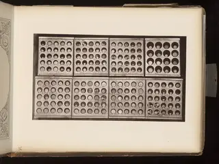 Eight wooden trays of small round coins in circular recesses, photographed from above, mounted on an album page.