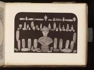 Many small vessels, cylindrical objects and a bust arranged in rows on a dark background