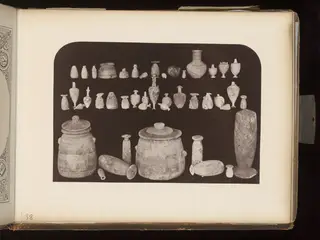 Rows of small vessels and juglets with two large lidded jars and a tall narrow vessel on a dark ground
