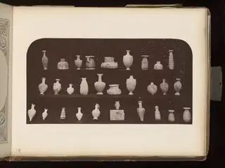 Small pale pottery vessels arranged on three dark shelves, with a small head on a block at lower right