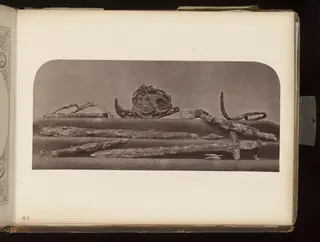 Corroded metal blades, a curved bit-like piece and a disc with a raised face, laid out on a cloth-covered shelf.