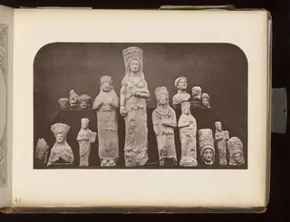 Pale statuettes and heads of figures with tall headdresses, arranged on dark boards against a dark background.