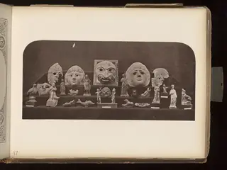 Masks, small figurines and animal figures arranged in rows on dark cloth shelves, mounted on an album page.