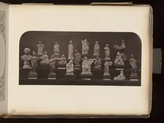 Rows of small figurines on stands, including a bust, draped standing figures and a cockerel, against a dark background