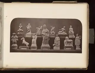 Fourteen small figurines of humans and animals arranged in two rows on wooden bases against a dark backdrop.