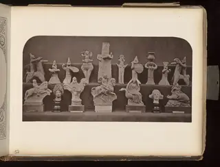 Two rows of small pale terracotta figurines of horses, riders, birds and figures arranged on shelves against a dark background.