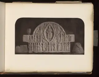 Arched carved stone relief panel with rows of small figures, flanked by two inscribed stone fragments on a dark background.