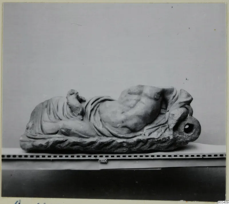 Reclining draped figure in marble with a broken head, with a ring-shaped loop at one end, shown beside a scale bar