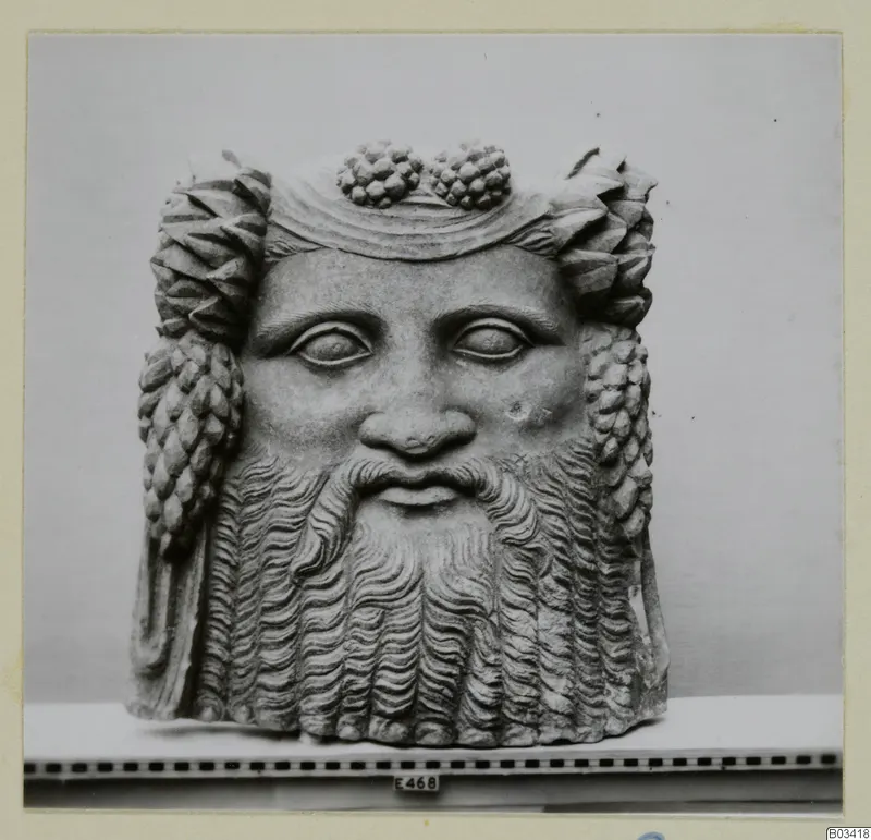Carved stone head of a bearded man, seen from the front, with clusters of grapes on top of the head