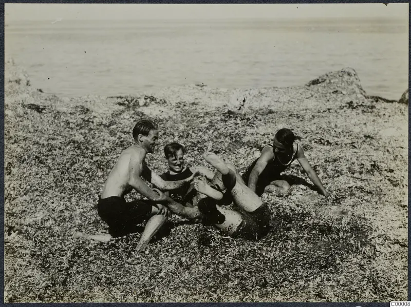 Four people playing and wrestling on a rocky, seaweed-covered shore beside the sea at Kyrenia.