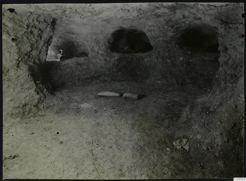 Tomb 313 a at Lapithos: interior of a rock-cut tomb chamber with arched niches, two stone slabs on the floor and a vessel in a niche