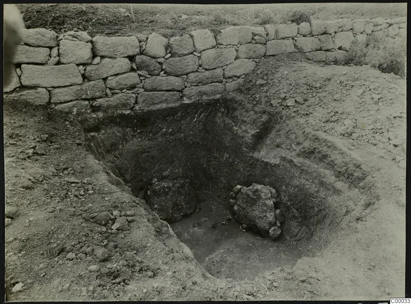 Excavated pit of Tomb 301 at Lapithos, with a dry-stone wall along its far edge and large rocks inside