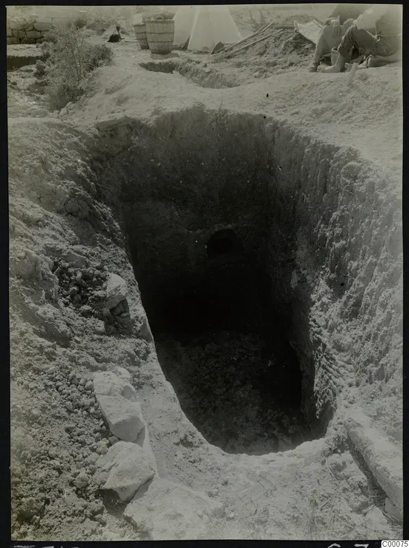 Tomb 313 at Lapithos: excavated rectangular shaft with a dark opening in the far wall, a kneeling worker and baskets beside it.