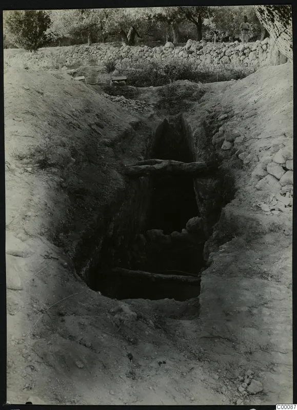 Tomb 411 at Lapithos during excavation, with a timber-braced cut in the ground and a standing man beyond.