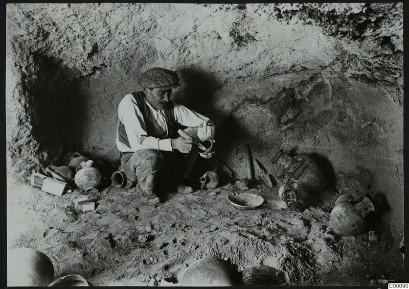 Man in a cap brushing pottery among vessels inside a rock-cut tomb chamber