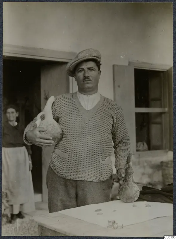 Man in a cap holding two pottery vessels, one in each hand, with a woman in a doorway behind him.