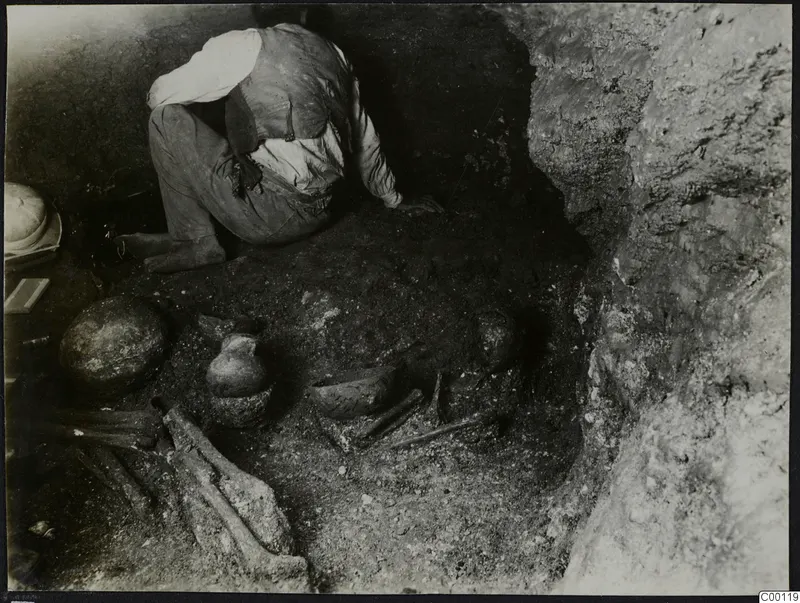 Tomb 306 c. at Lapithos: a man kneels among pottery vessels and bones in an excavated tomb chamber.