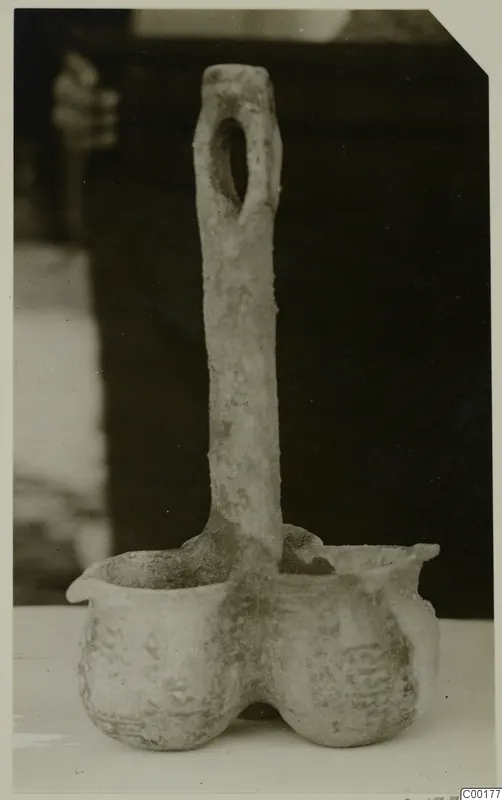Pottery vessel with two deep rounded bowls joined at the base and a tall flat handle with an oval hole at the top