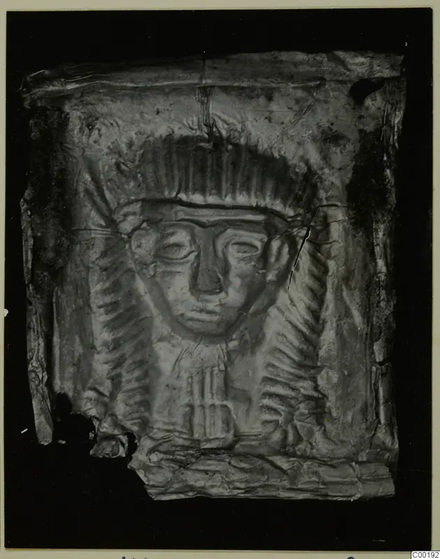 Embossed metal plaque with a raised face framed by a ridged headdress; the sheet is crumpled and torn at the edges