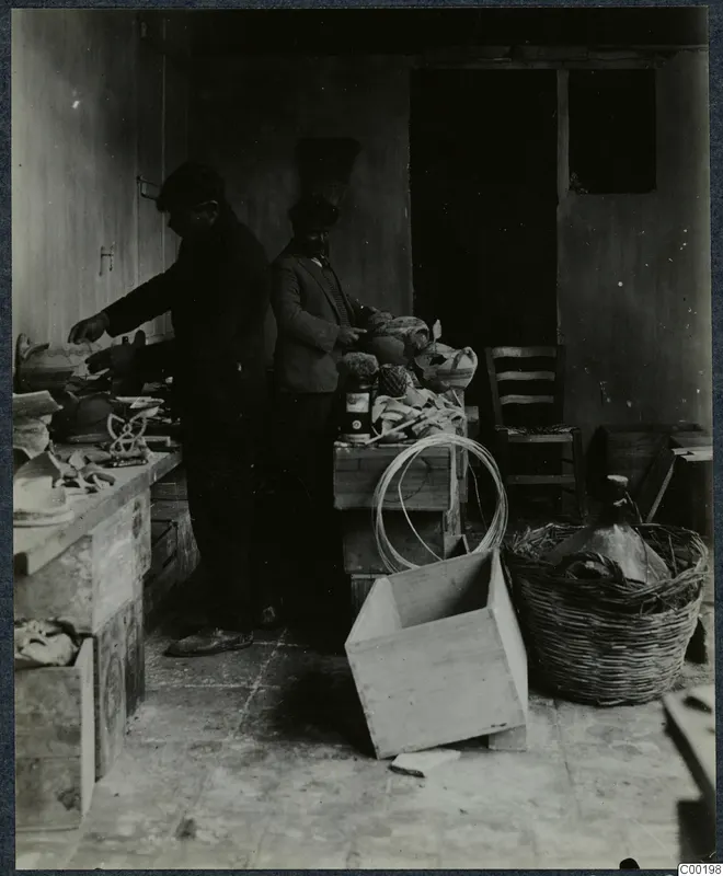 Two men in caps working among pottery vessels, crates and a wicker basket in a bare storeroom