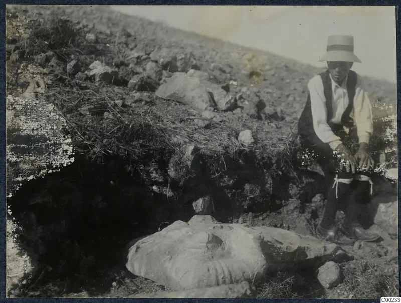 Carved stone fragment lying in an excavated hillside at Vouni, with a seated person in a hat beside it