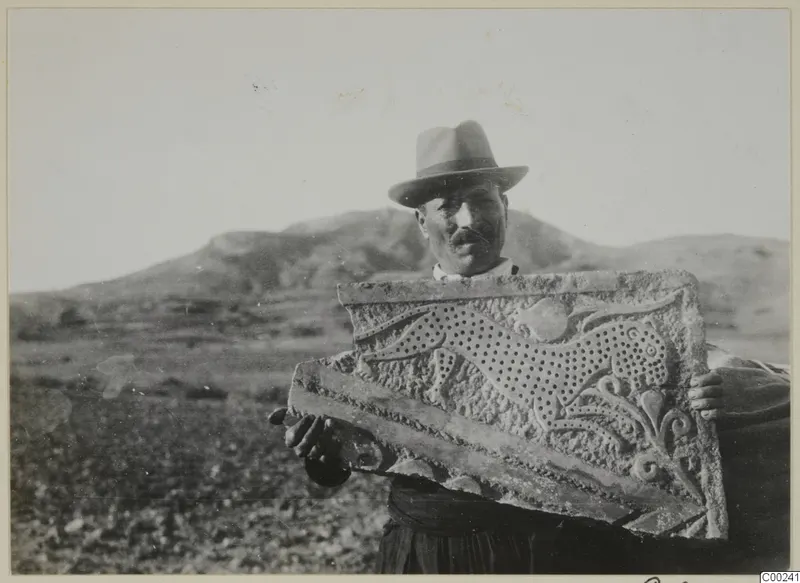 Man in a hat holding a carved stone relief panel with a feline figure and leafy ornament, low hills behind him