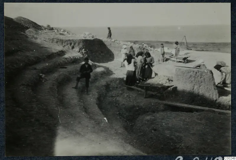 Excavation at Soli theatre with workers, a wheelbarrow and cut stone blocks, the sea on the horizon
