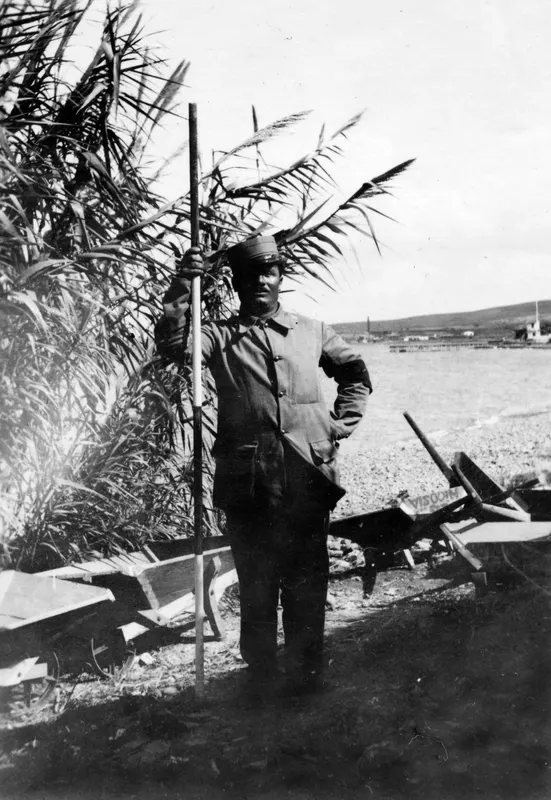 Toulis Souidos holding a measuring staff on a pebble beach at Soli, with reeds and wheelbarrows behind him.