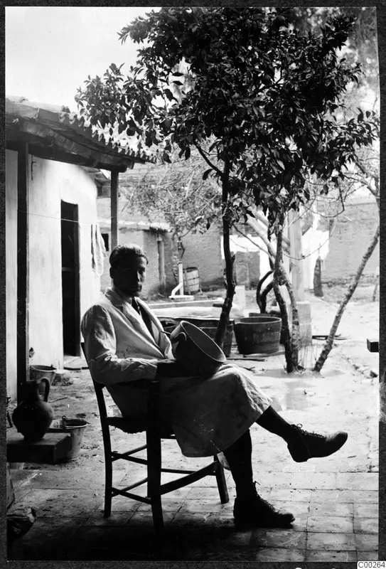 Alfred Westholm seated on a wooden chair in a courtyard, holding a wide vessel on his lap.