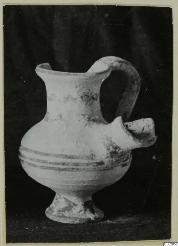 Pale clay vessel with a side spout, a loop handle and dark painted bands, photographed against a dark backdrop
