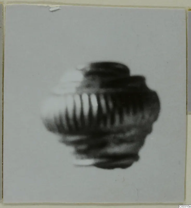 Blurred photograph of a single ribbed, rounded object, possibly a bead or pendant, on a plain grey background