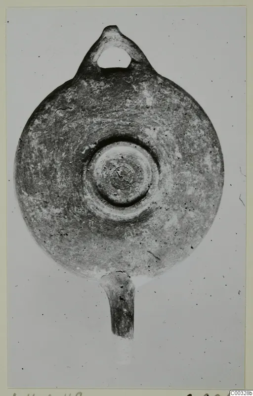 Plain clay oil lamp seen from above, with a loop handle at the top and a nozzle at the bottom.