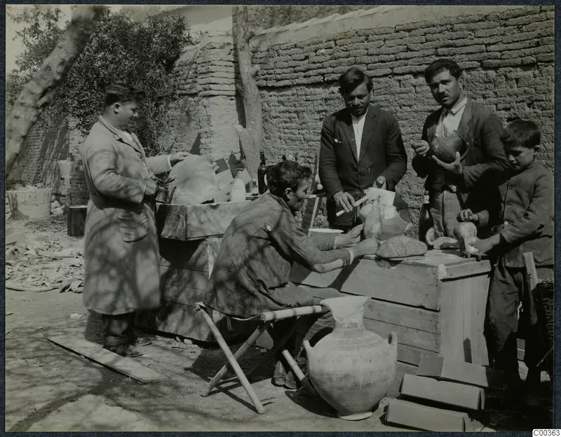 Four men and a child examine pottery on wooden boxes used as tables in an open yard by a mud-brick wall