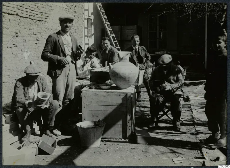 Six men and a child gathered around wooden crates with a large pale jar, in a sunlit courtyard beside a mud-brick wall