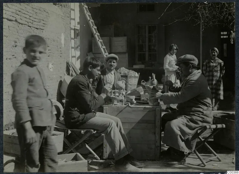 Three men at a wooden crate examining pottery outside a house, with a child and two women nearby