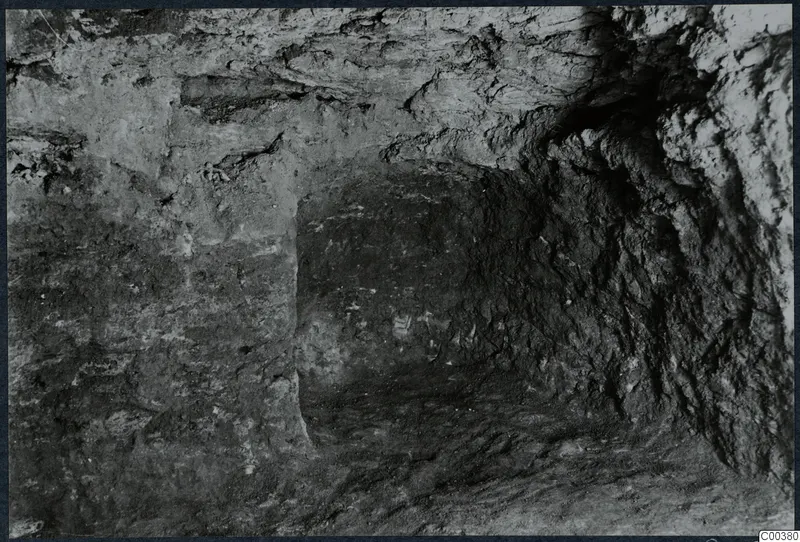 Interior of Tomb 13 at Milia during excavation, with rough rock walls and an uneven floor.
