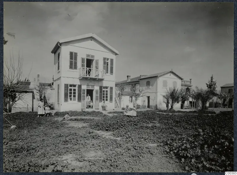 Two-storey white house with a first-floor balcony and people at its doorway, in a garden in Nicosia
