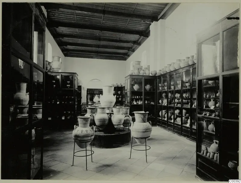 Museum gallery with large painted pottery jars on metal stands and many vessels in glass cabinets