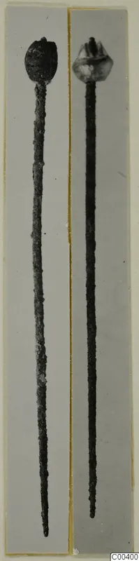 Two long pins with decorated heads, one dark and one lighter, photographed side by side on grey mounts.