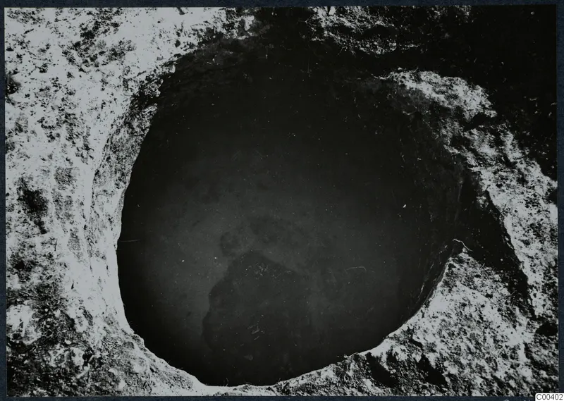 Excavated tomb at Milia seen from above, a round opening in pale soil with a dark mass of stones at the bottom