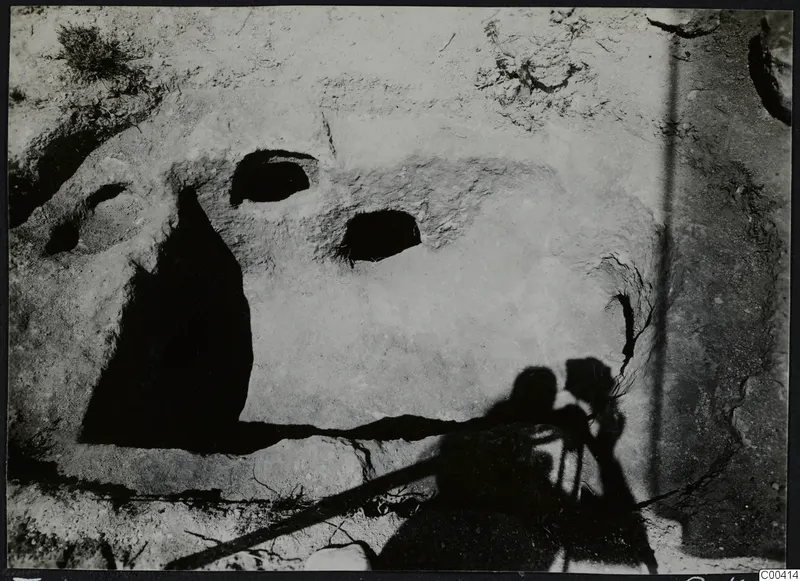 Tomb 307 at Vrysi tou Barba, Lapithos, during excavation, showing two openings in a pale rock surface with grass at the edge.