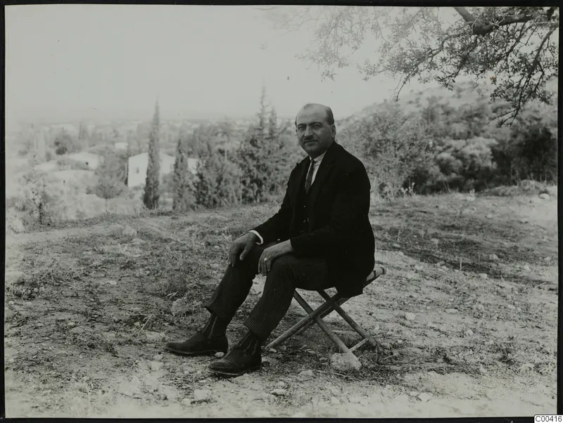 Man in a dark suit seated on a folding stool outdoors, with trees and a white house behind him.