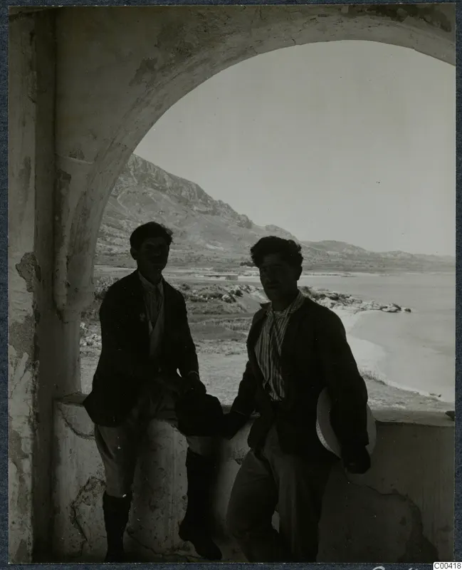Two men standing in an arched opening, with a bay of sea and a mountain ridge behind them.