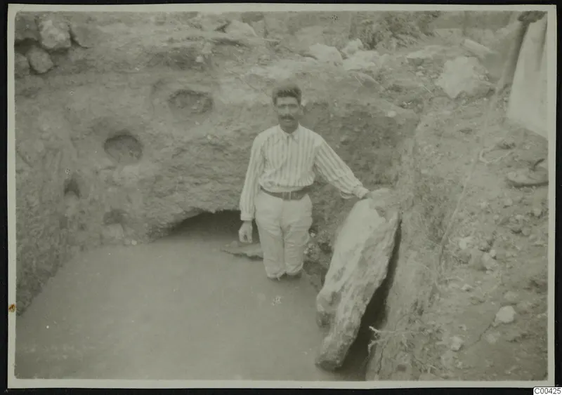 Man standing in water at the bottom of an excavation trench, beside a large pale stone slab