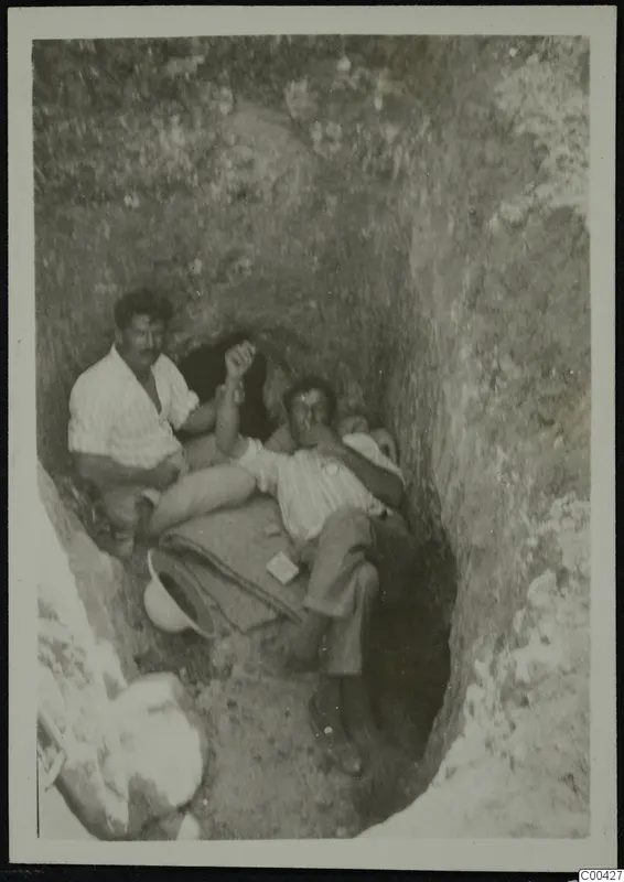 Two men resting in an excavation cutting at Lapithos, with a white helmet on the ground beside them.