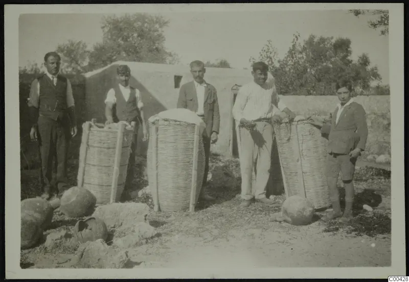 Five men and boys stand beside large wicker baskets and jars in front of a whitewashed building at Lapithos.