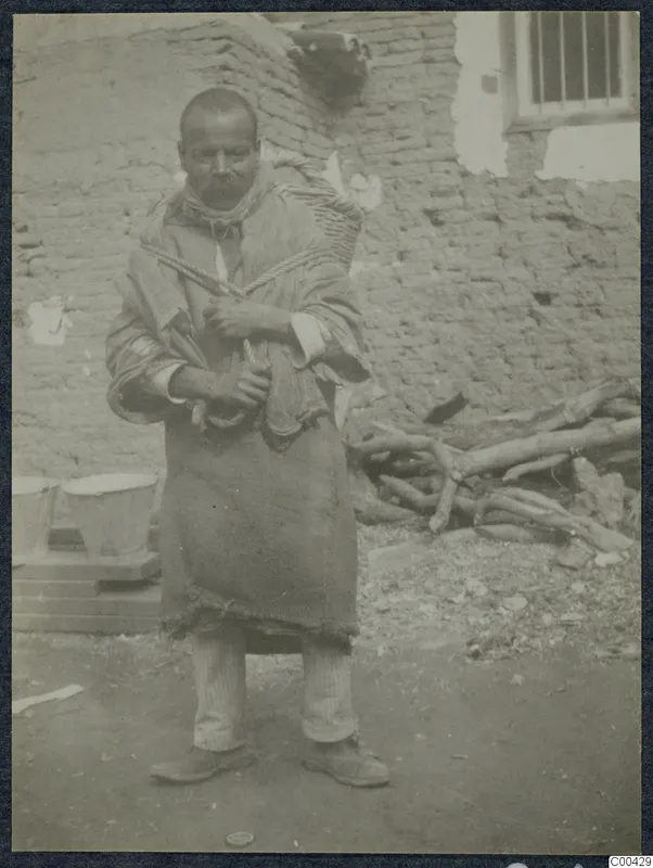 Man with a moustache carrying a basket on his back, standing in front of a mud-brick wall