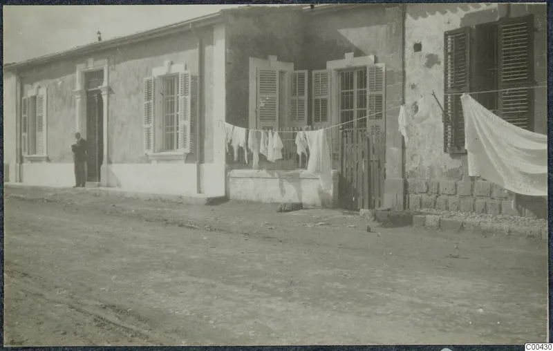 Single-storey building with shuttered windows, a standing man at the doorway and laundry hung on a line in front.